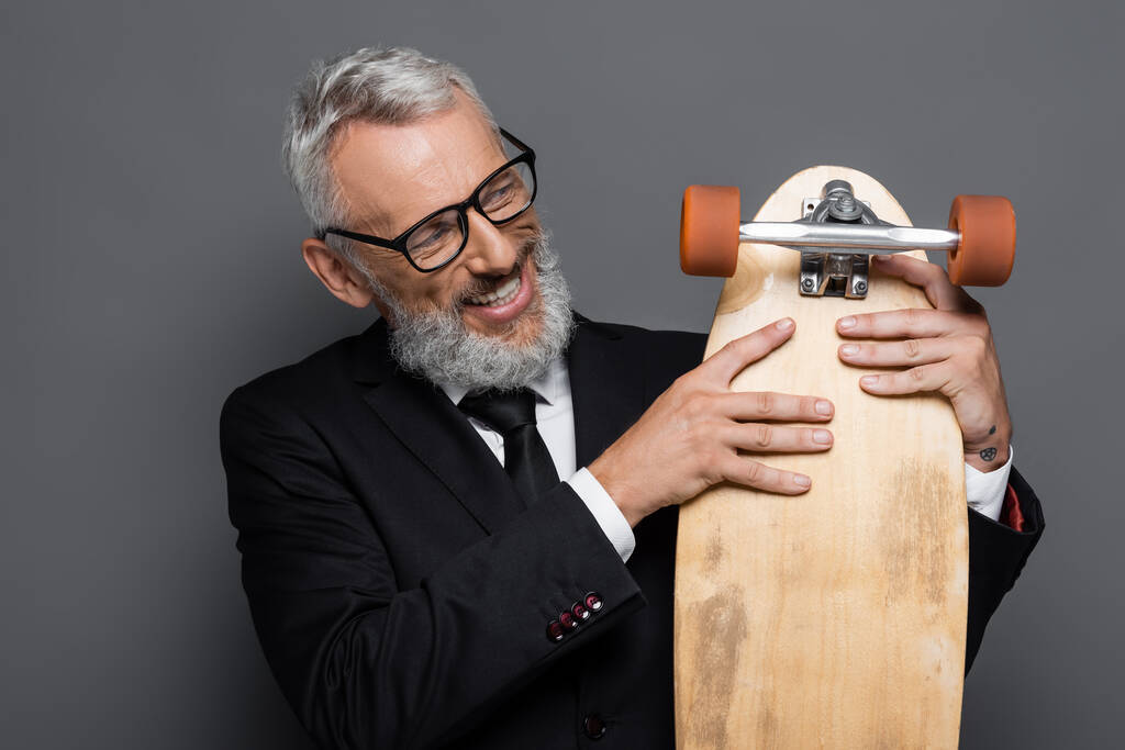 glücklicher und reifer Geschäftsmann in Brille mit Longboard auf grau  - Foto, Bild