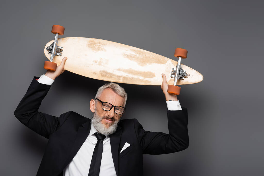 homme d'affaires joyeux et mature en costume et lunettes tenant longboard au-dessus de la tête sur gris  - Photo, image