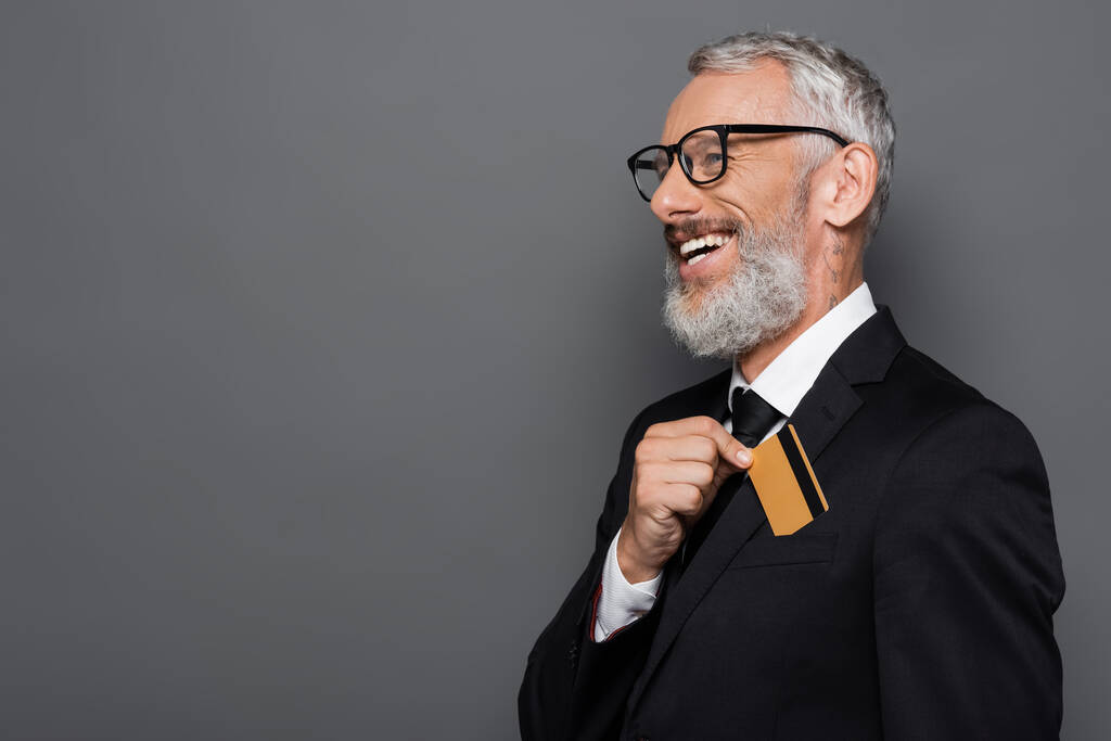 feliz hombre de negocios de mediana edad en traje y gafas de poner la tarjeta de crédito en el bolsillo aislado en gris - Foto, imagen