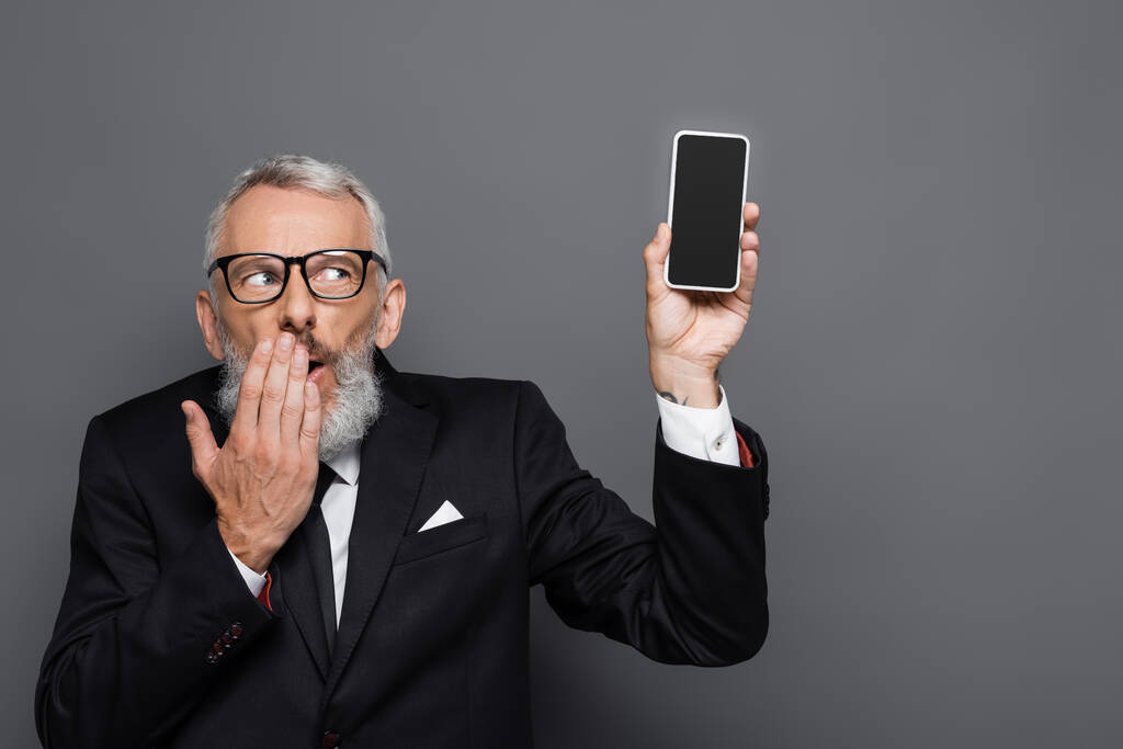 středního věku podnikatel drží mobilní telefon s prázdnou obrazovkou a zakrývá ústa na šedé - Fotografie, Obrázek