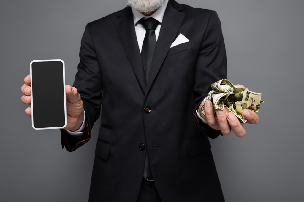abgeschnittene Ansicht von Geschäftsmann mittleren Alters mit Dollarnoten und Smartphone mit leerem Bildschirm isoliert auf grau - Foto, Bild