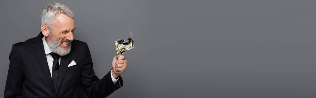 szczęśliwy przedsiębiorca w średnim wieku trzyma płonące banknoty dolarowe odizolowane na szarym, baner - Zdjęcie, obraz
