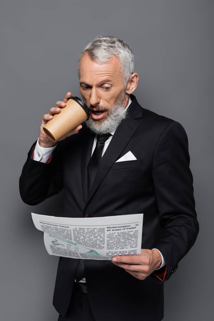 šokovaný podnikatel středního věku drží papírový pohár a čtení novin na šedé - Fotografie, Obrázek