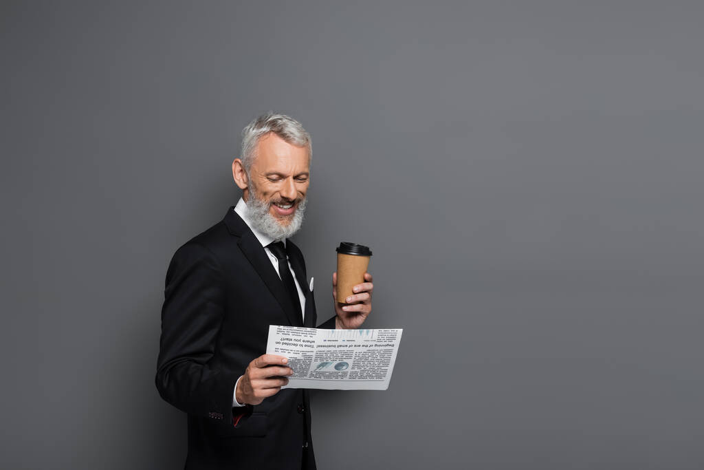 radostný podnikatel středního věku drží papírový šálek a čte noviny na šedé - Fotografie, Obrázek