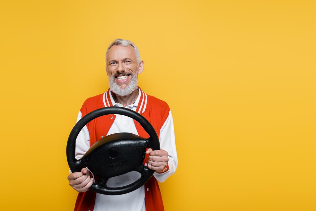 alegre homem de meia idade em jaqueta bombardeiro segurando volante enquanto imitando condução isolada no amarelo - Foto, Imagem