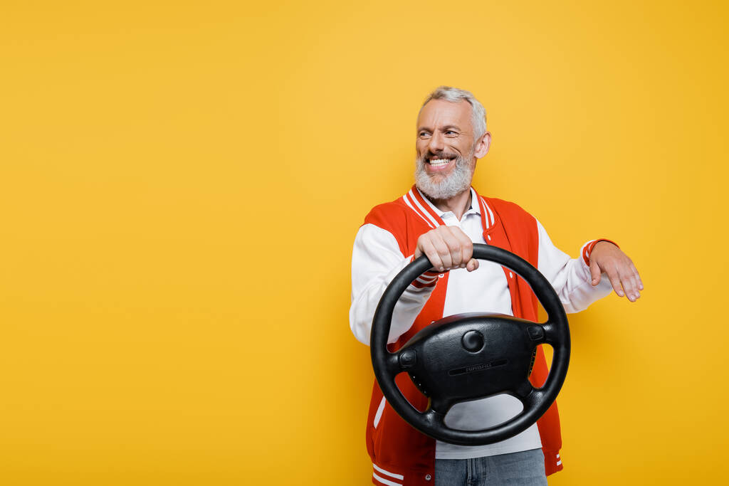 szczęśliwy mężczyzna w średnim wieku w kurtce bombowej trzymając kierownicę podczas gestów odizolowanych na żółto - Zdjęcie, obraz