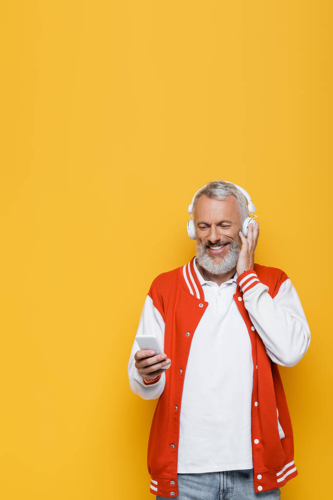 positive middle aged man in wireless headphones holding cellphone and listening music isolated on yellow  - Photo, Image