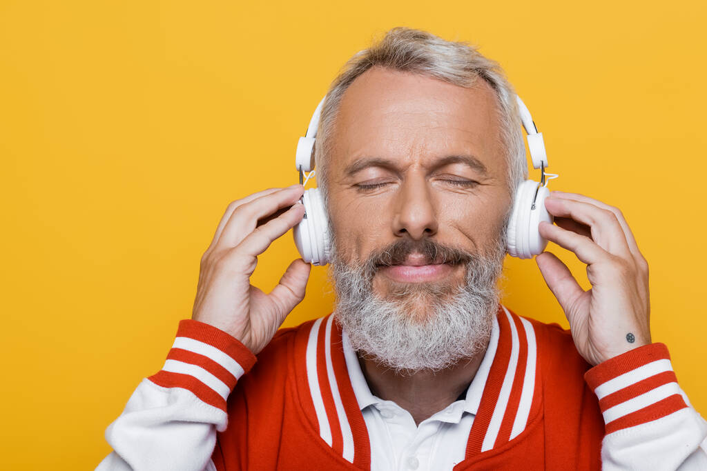 positive middle aged man in wireless headphones listening music isolated on yellow  - Photo, Image