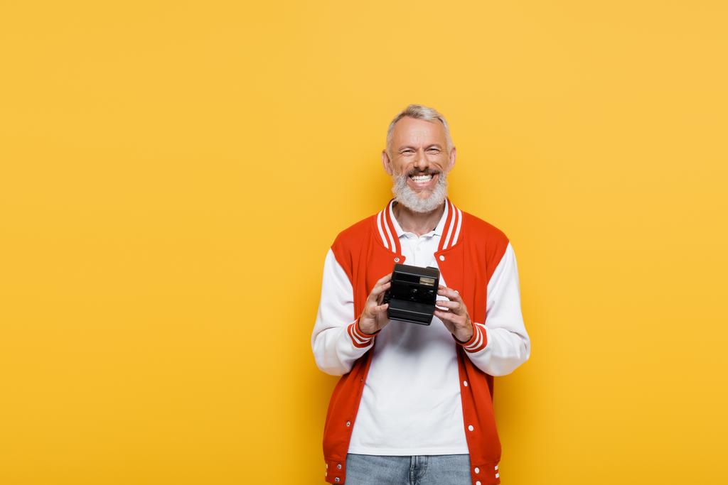 joyful middle aged man in bomber jacket holding black vintage camera isolated on yellow - Photo, Image
