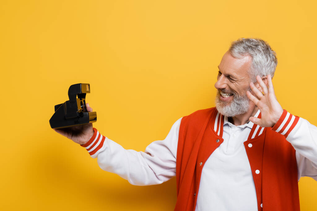 szczęśliwy mężczyzna w średnim wieku w kurtce bomber biorąc selfie na czarny vintage aparat na żółty - Zdjęcie, obraz