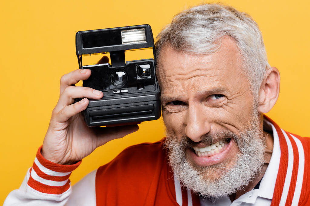 middle aged man in bomber jacket holding black vintage camera and grinning isolated on yellow - Photo, Image