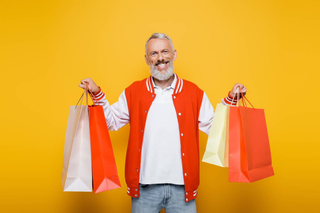 glücklicher Mann mittleren Alters in Bomberjacke mit gelben Einkaufstaschen - Foto, Bild