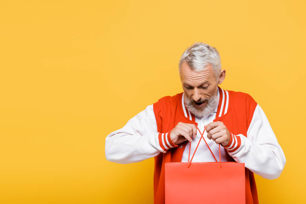 изумленный мужчина средних лет в куртке подрывника держит сумку с покупками на желтом - Фото, изображение