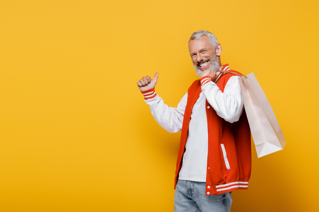 feliz hombre de mediana edad en chaqueta de bombardero sosteniendo bolsa de compras y mostrando el pulgar hacia arriba en amarillo - Foto, Imagen