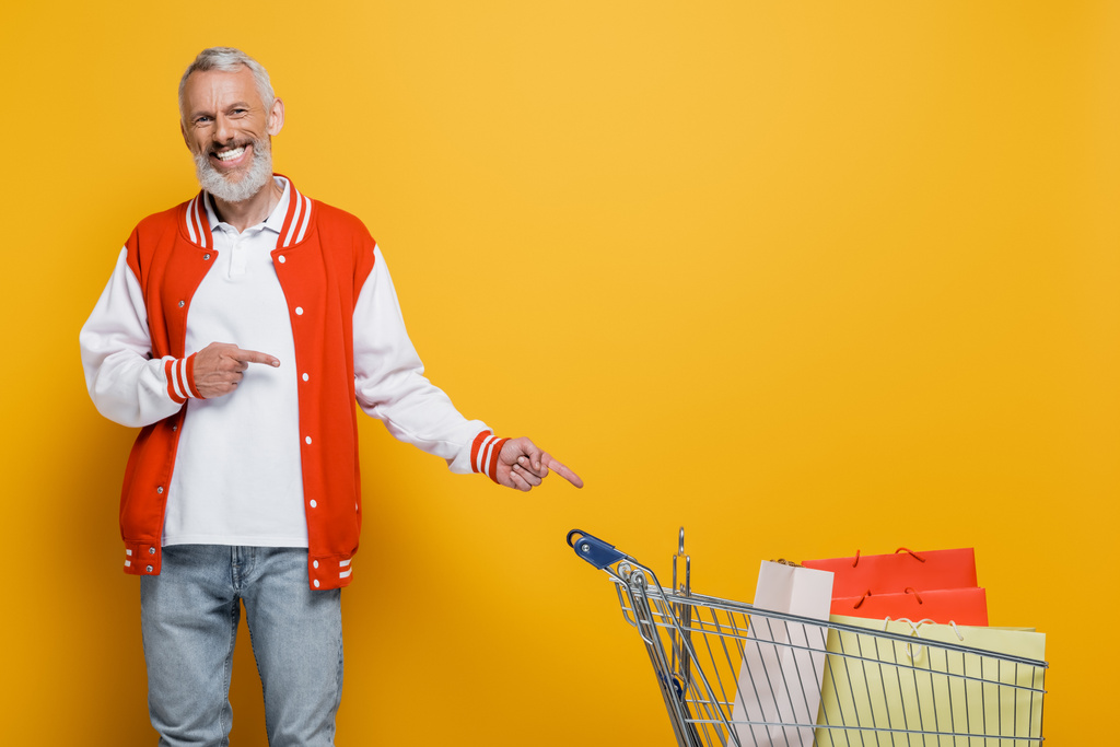 zadowolony mężczyzna w średnim wieku w kurtce bombowej wskazując na wózek z torbami na zakupy na żółty - Zdjęcie, obraz