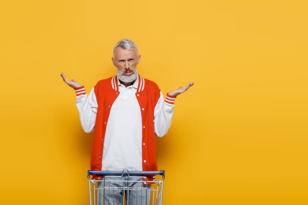 confuso hombre de mediana edad en chaqueta de bombardero mostrando gesto encogiéndose de hombros cerca de la cesta de la compra vacía en amarillo - Foto, Imagen