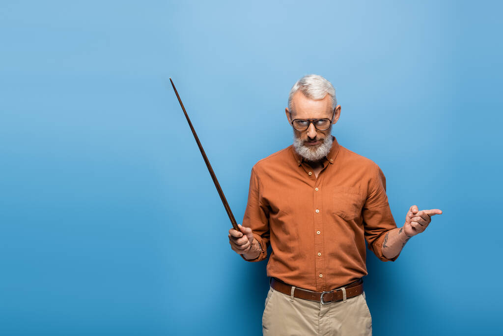 tattooed middle aged teacher in glasses holding pointer stick and pointing away on blue - Photo, Image