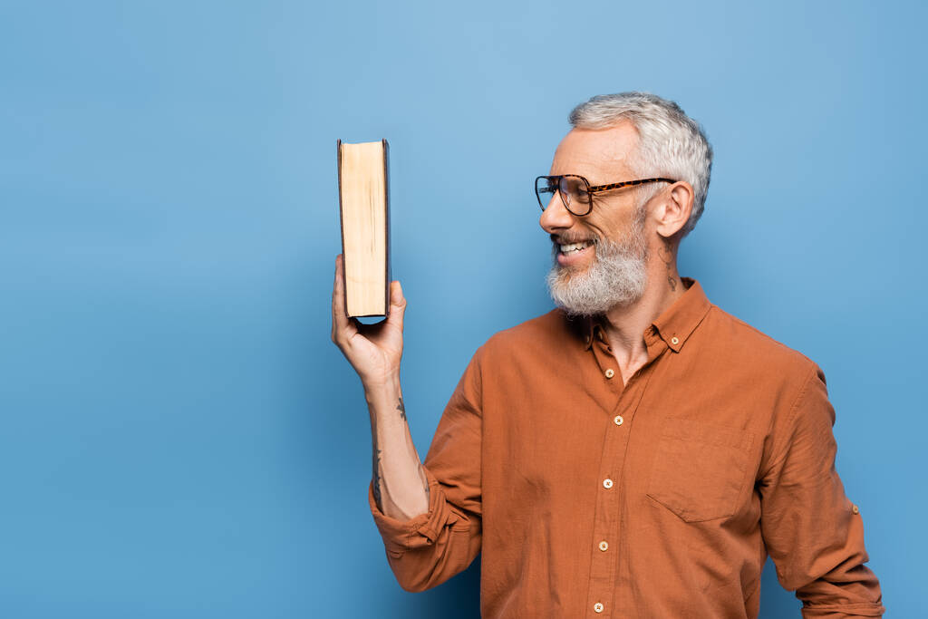 Dövmeli orta yaşlı gözlüklü öğretmen gülümsüyor ve mavi üzerine kitap tutuyor. - Fotoğraf, Görsel