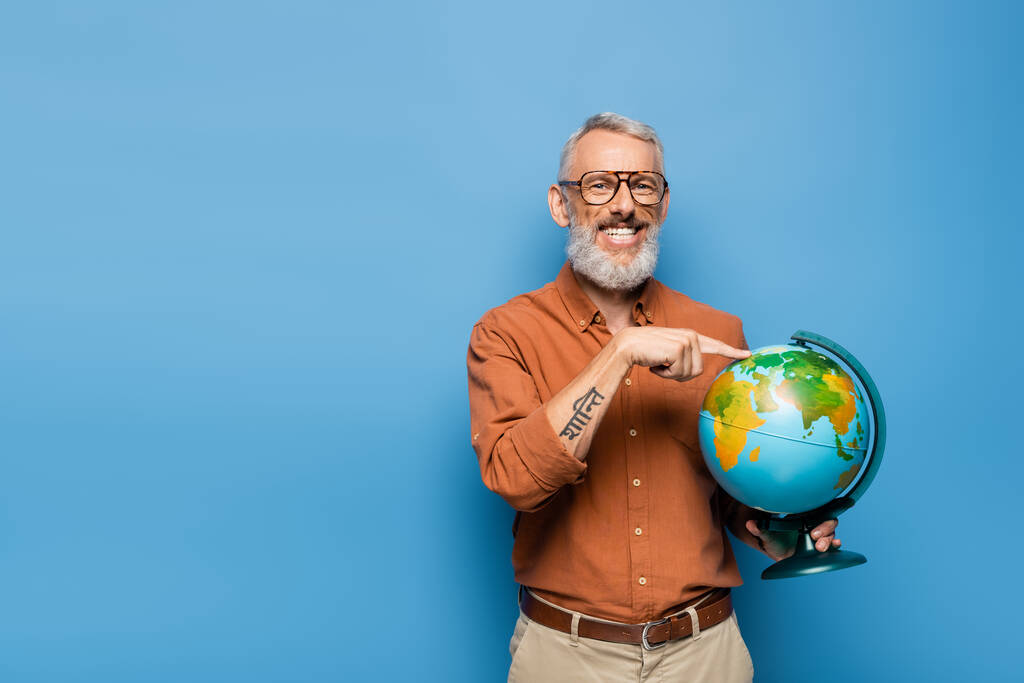 щасливий вчитель середнього віку в окулярах, вказуючи пальцем на глобус на синьому
 - Фото, зображення
