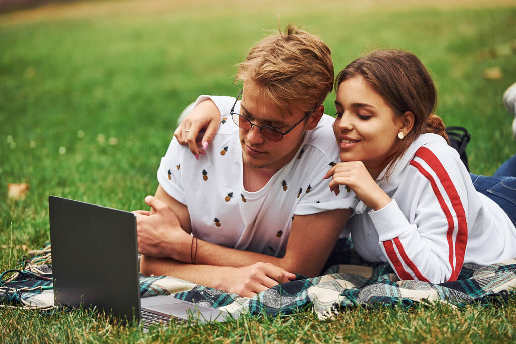 Młoda para siedzi na zielonej trawie na boisku z laptopem. - Zdjęcie, obraz