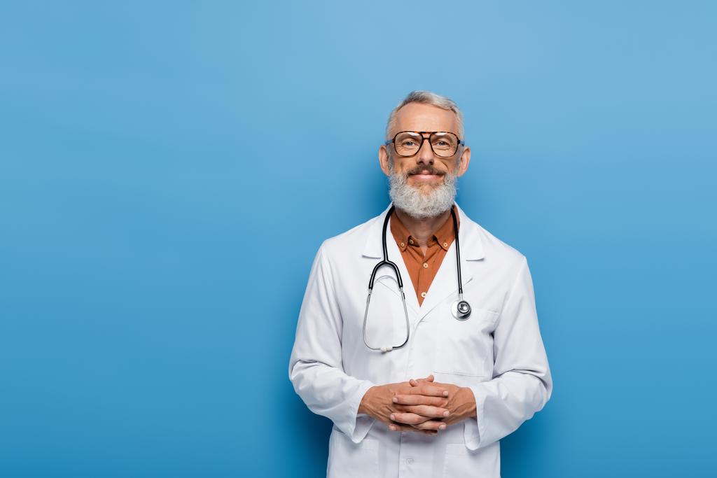 gelukkig middelbare leeftijd arts in bril staan met gebalde handen op blauw - Foto, afbeelding