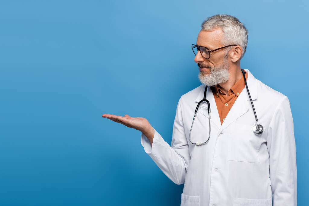 médico barbudo de mediana edad en gafas y abrigo blanco apuntando con la mano sobre azul - Foto, imagen