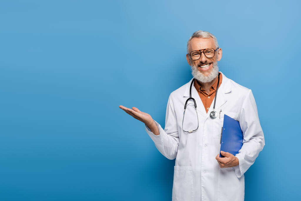 gelukkige dokter van middelbare leeftijd in witte jas en bril met klembord en wijzend met de hand op blauw - Foto, afbeelding