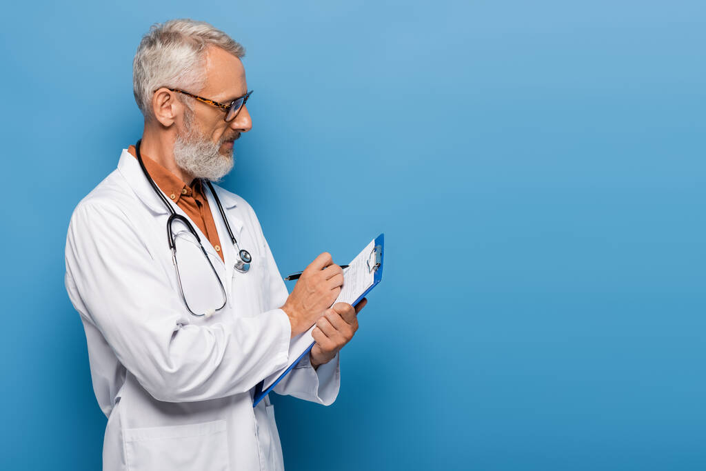 bärtiger Arzt mittleren Alters in weißem Mantel und Brille, der Rezepte auf Klemmbrett auf blau schreibt - Foto, Bild