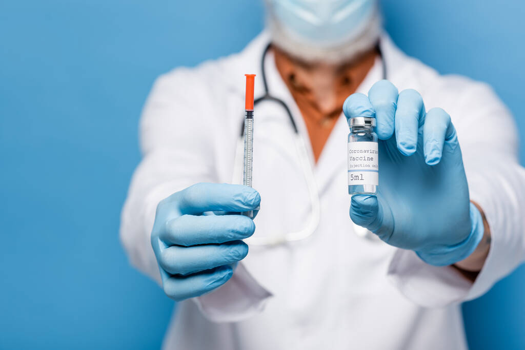обрезанный взгляд врача в латексных перчатках, держащих флакон с коронавирусом вакцины и шприца изолированы на голубом - Фото, изображение