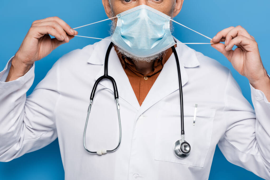 cropped view of middle aged doctor in white coat adjusting medical mask isolated on blue  - Photo, Image