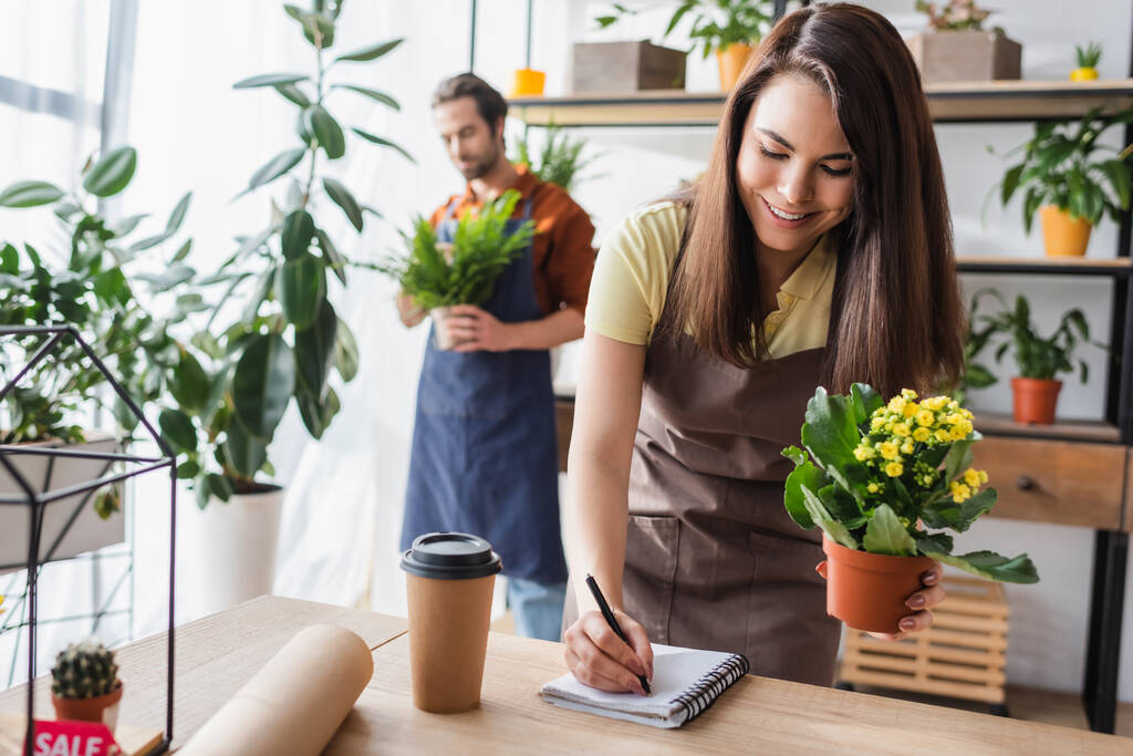 Young seller holding plant and writing on notebook near blurred colleague in flower shop  - Photo, Image