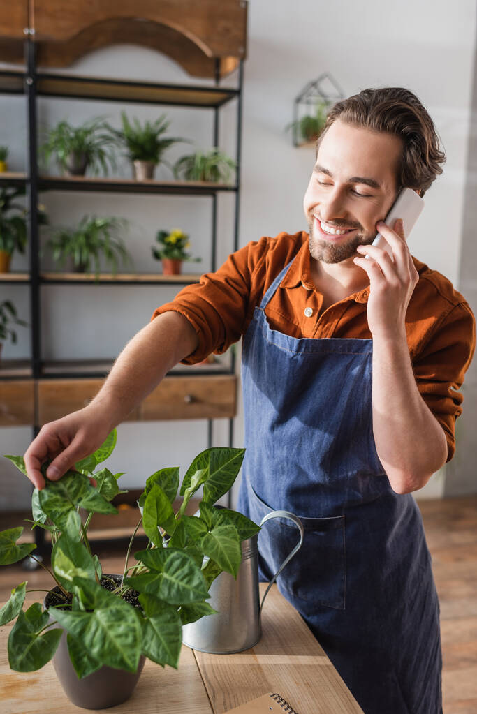 Smiling florist in apron talking on smartphone near plant and watering can in flower shop  - Photo, Image
