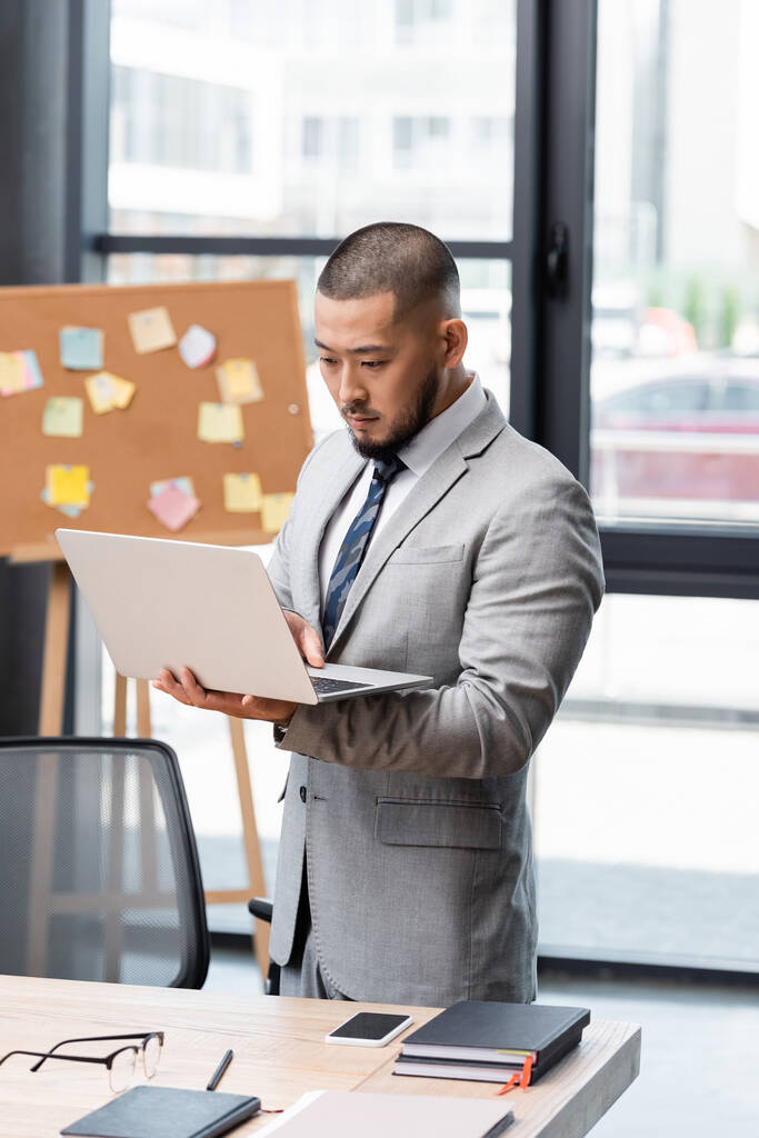 азіатський бізнесмен у формальному одязі, що стоїть з ноутбуком біля робочого столу в офісі
 - Фото, зображення