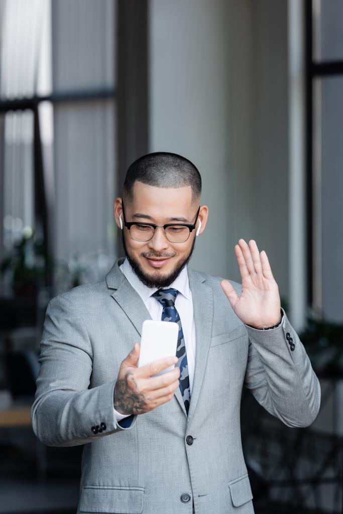 sonriente asiático hombre de negocios en auriculares agitando la mano durante la videollamada en el teléfono inteligente - Foto, Imagen