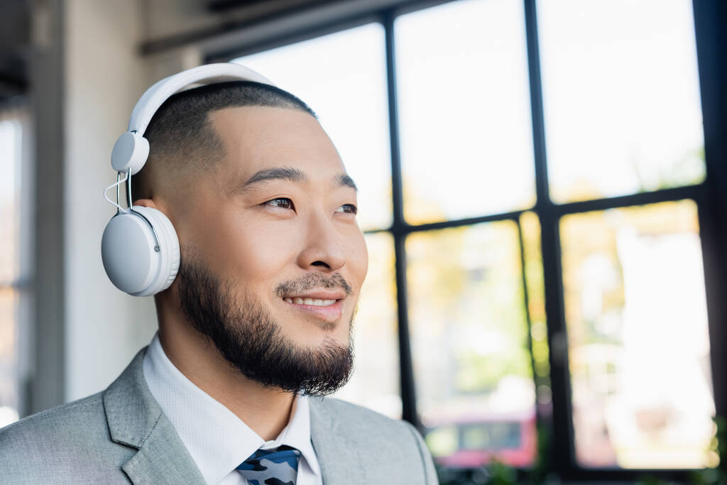 gelukkig aziatische zakenman in draadloze hoofdtelefoon op zoek weg in kantoor - Foto, afbeelding