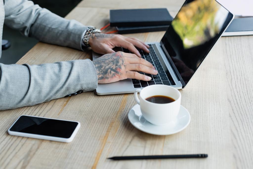 vista ritagliata di uomo d'affari tatuato digitando sul computer portatile vicino alla tazza di caffè e al cellulare con schermo vuoto - Foto, immagini