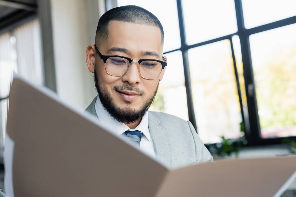 smiling asian businessman looking at documents in blurred folder in office - Photo, Image