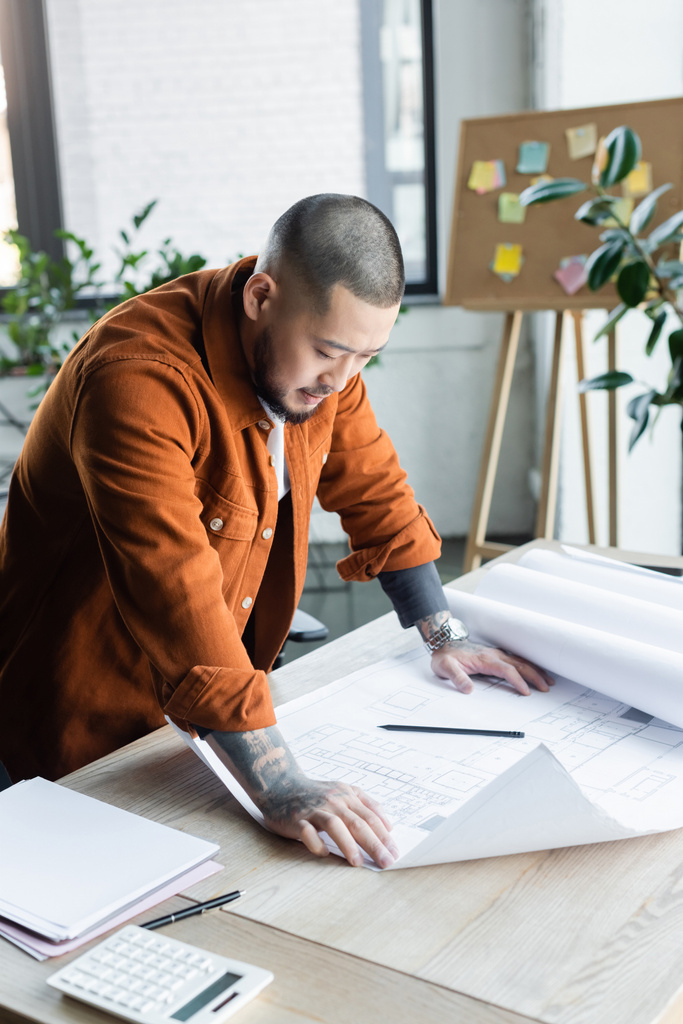 asiatico ingegnere guardando architettonico piano mentre in piedi a scrivania in ufficio - Foto, immagini