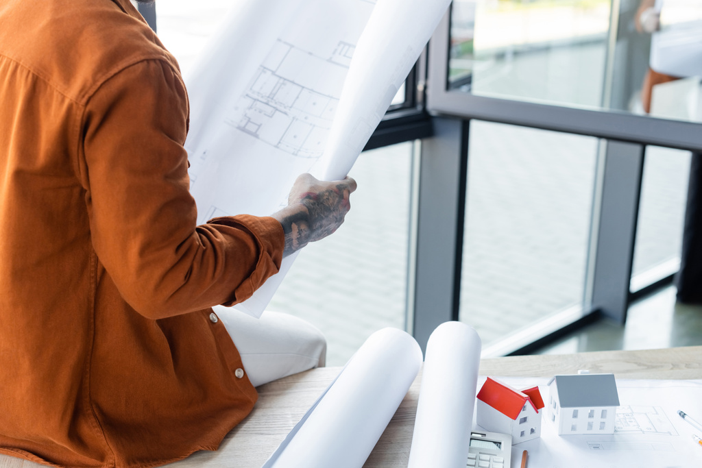 обрезанный вид архитектора, держащего план строительства, сидя на столе рядом с чертежами и моделями домов  - Фото, изображение