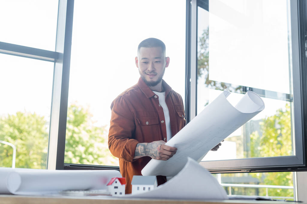 usmívající se asijský architekt při pohledu na rozmazané modely domu při držení náčrt v kanceláři - Fotografie, Obrázek
