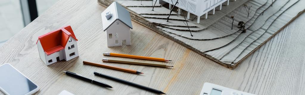 high angle view of pencils, house models and smartphone on wooden desk, banner - Photo, Image