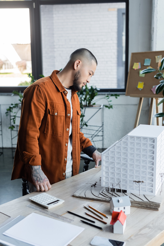 Ασιάτης αρχιτέκτονας με τατουάζ που στέκεται στο γραφείο εργασίας και κοιτάζει το μοντέλο του σπιτιού στο γραφείο - Φωτογραφία, εικόνα