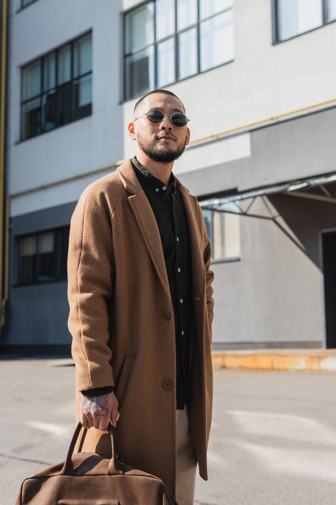 stylish asian man in sunglasses and beige coat standing near blurred building outdoors - Photo, Image