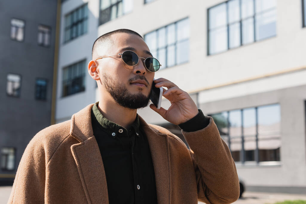 asian man in coat and sunglasses talking on mobile phone on urban street - Photo, Image