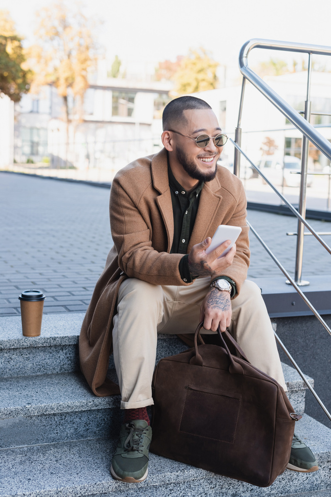 šťastný a stylový asijský muž s mobilním telefonem sedí na schodech v blízkosti papírového kelímku venku - Fotografie, Obrázek