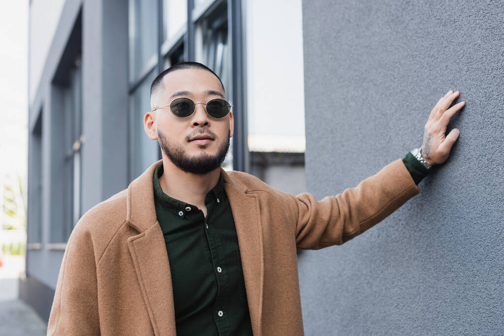 asian man in sunglasses and trendy autumn coat touching grey wall of building outside - Photo, Image