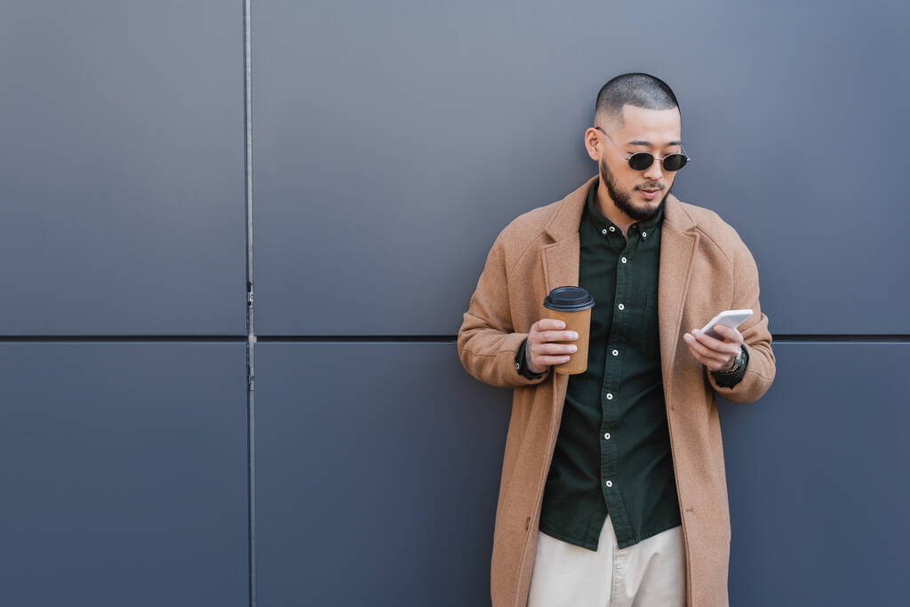 modny Azji człowiek z na wynos drink wiadomości na telefon komórkowy w pobliżu szary ściana - Zdjęcie, obraz