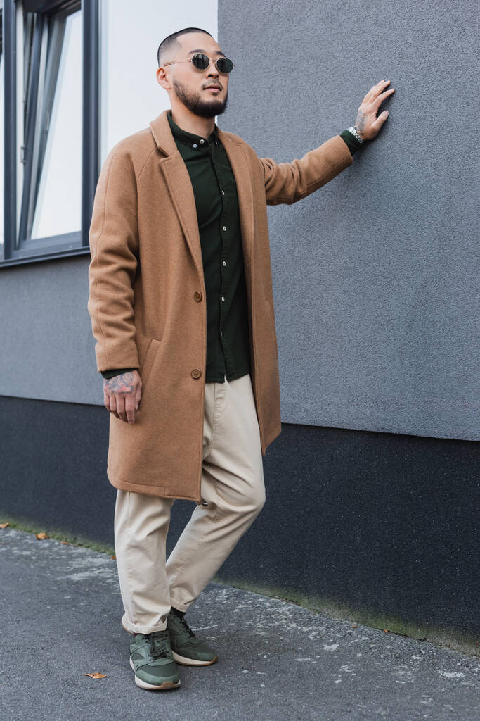 ásia homem no na moda casaco e óculos de sol inclinado no cinza edifício no urbano rua - Foto, Imagem