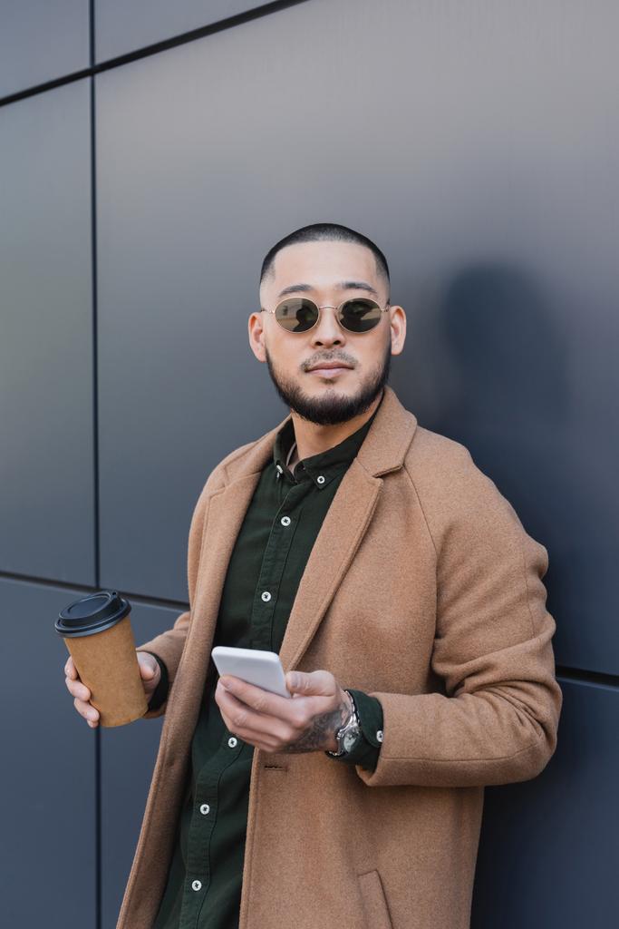 asiático hombre en moda otoño traje celebración takeaway beber y móvil cerca gris pared - Foto, imagen