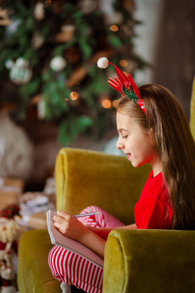 かわいい白人の女の子が左手でクリスマスツリーの近くにサンタクロースに手紙を書きます。幸せな子供時代、奇跡と欲望の時。メリークリスマス. - 写真・画像